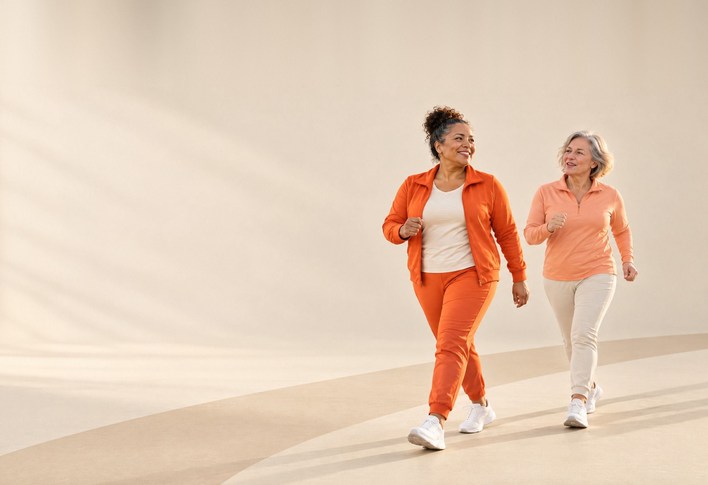 Two friends in coordinated orange and apricot walking gear stride across a warm sand-coloured studio set, laughing.