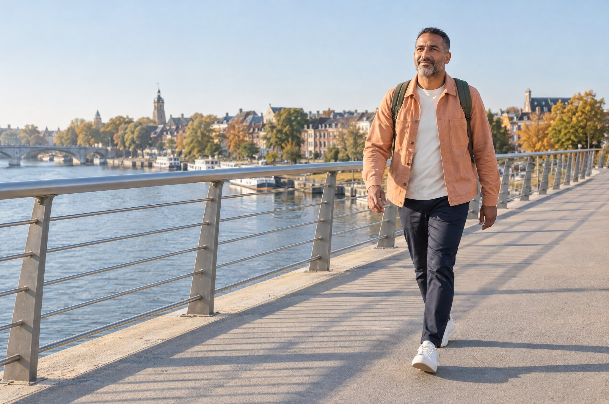 A man with a small backpack walks across a city footbridge over a river on a sunny autumn morning.