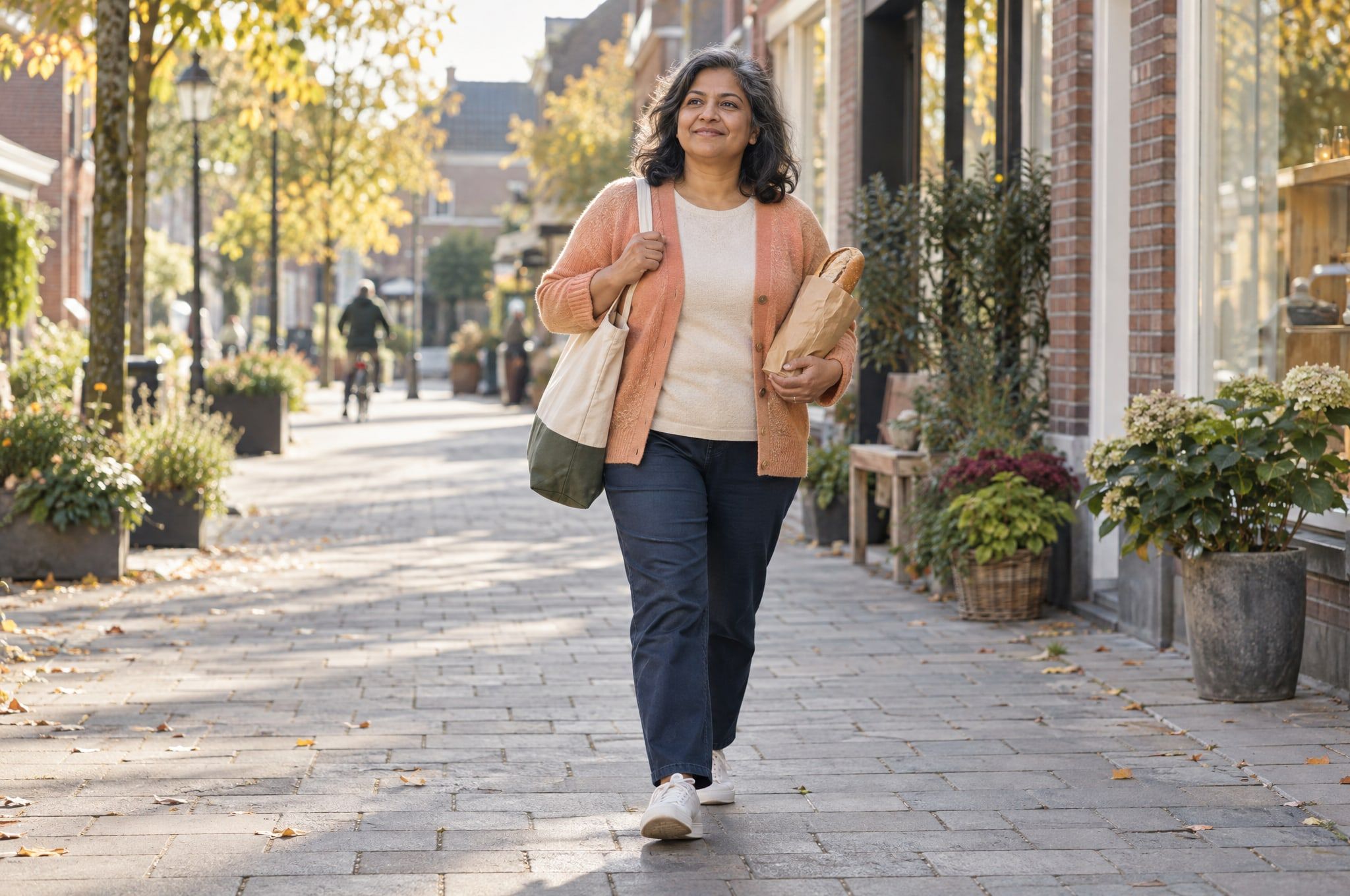 A woman in an apricot cardigan walks down a sunny shopping street with a tote bag and a loaf of bread.